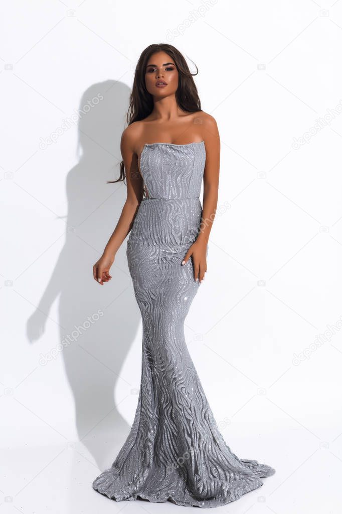 Picture of beautiful slim model in silver long dress on white background. Brunette volume hairstyle and modern makeup.