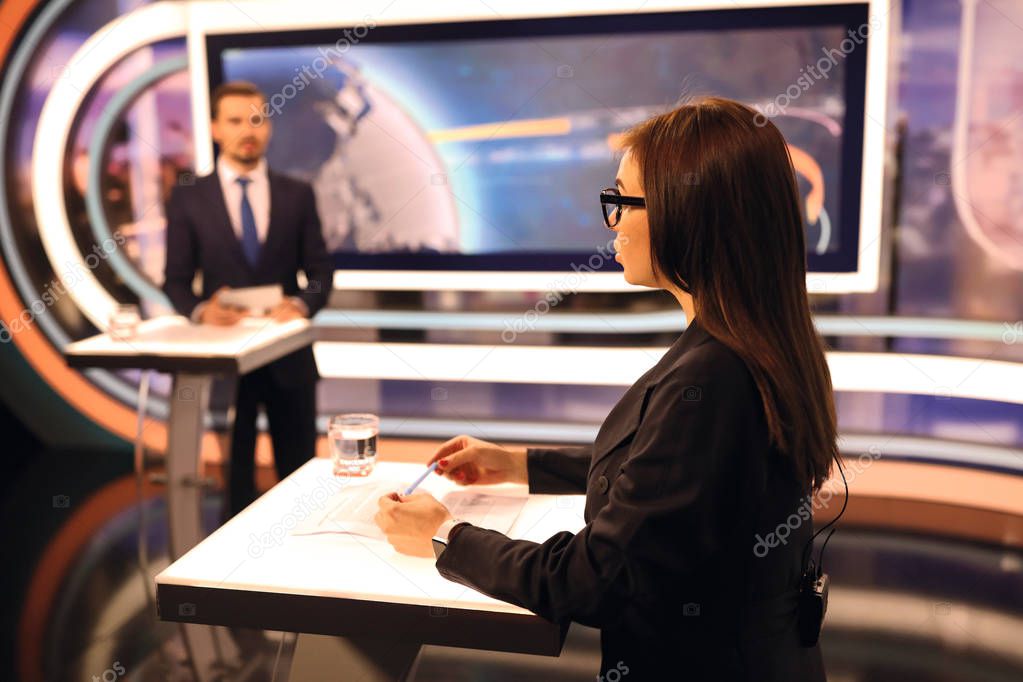 Woman in glasses and man in suit standing in studio, making a TV news show. A glass of water and paper leaf on the table. Big screen on the background.