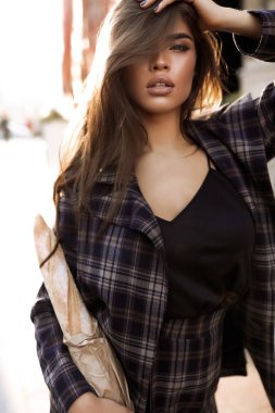 Güzel model sokakta baget ile. Moda açık ateş. Seksi ince gövde, çıplak makyaj ile güzel yüzü. Şık uzun kahverengi saçlı. Siyah gömlek ve gri kareli ceket giyiyor. Güneş ışınları arkasında