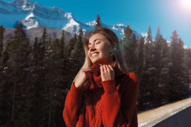 Gülen gözlerin kapalı al kış kızla sunbathes. Saf mavi gökyüzü ve arka planda büyük dağlar. Düzgün vücut üzerinde sıcak kırmızı örgü kazak. Alaska.White dişler, ihale cilt soğuk kış moda