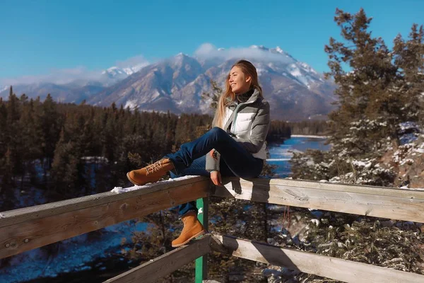かなりの女性の冬のファッションと快適な服を着て寒さと雪に覆われたカナダを旅行します 遠くを見て 彼女の柔らかい顔に太陽の光をキャッチ 木製フェンスの上に座っています 絵のような — ストック写真