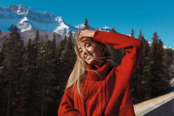 快乐漂亮的女士穿着红色毛衣站在户外 欣赏她周围的自然景观 摸着金发 面带微笑 没有化妆 自然之美 密集的森林和高山在背景 — 图库照片