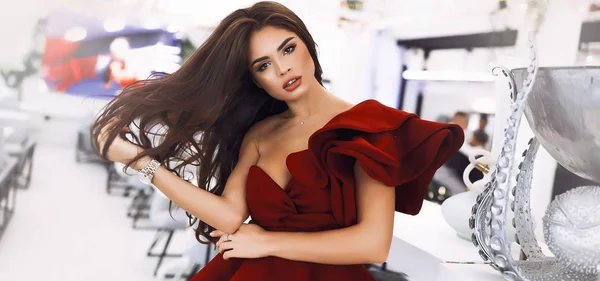 Passion lady throwing her long brunette hair.Bright fashion makeup and big lips. Wearing burgundy party dress with sleeve and decollete.Hand on the waist. Standing near the bar counter and silver vase