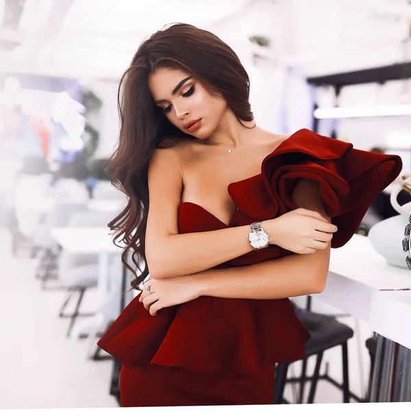 Looking down lady in burgundy evening dress and makeup standing near the bar counter, holding herself. Accessories on hand and neck. Long brunette hair falling down on back. Pretty face with big lips