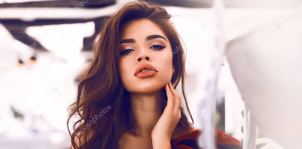 Amazing brunette model with beautiful makeup on the face, big lips and soft skin. Wearing burgundy dress with decollete and open shoulder. Stylish curly hair lying on the shoulder, hand touching neck