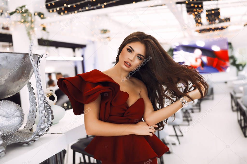 Amazing lady in red party dress with volume sleeve and decollete standing in luxury restaurant. Touching her long dark hair, head tilted on side. Fashion shooting for modern collection. Cozy interior