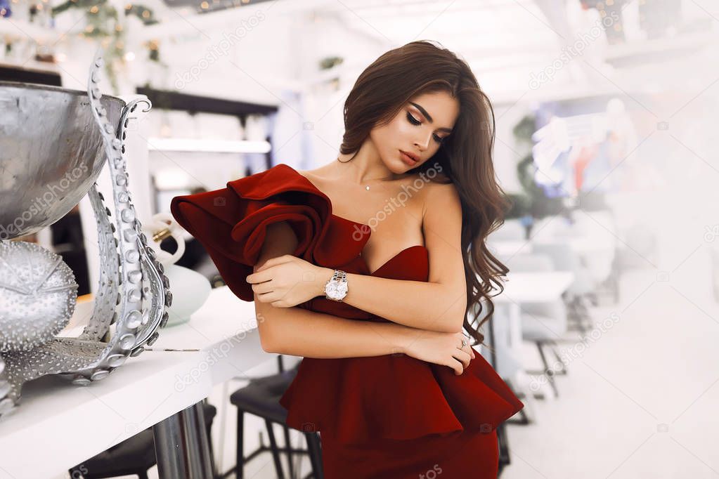 Tender female lean on her shoulder, looking down, holding herself. Watches on a hand, shining accessories. Long curly hairstyle and bright evening makeup. Cocktail red dress on slim body. Silver vase