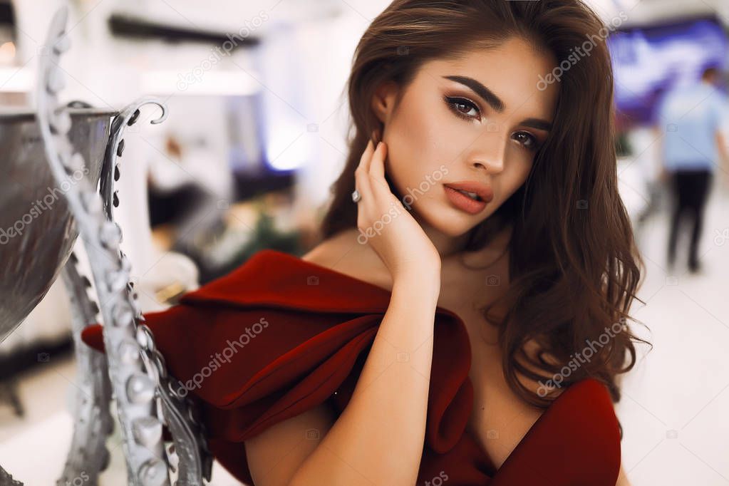 Lovely female looking forward, beautiful eyes, big lips and nude makeup. Hand on the neck, accessories. Dark brown hair lying on the chest. Standing near the silver designer vase. Wearing red dress