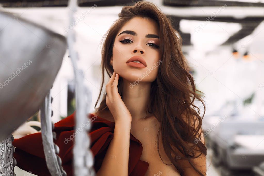 Amazing brunette model with beautiful makeup on the face, big lips and soft skin. Wearing burgundy dress with decollete and open shoulder. Stylish curly hair lying on the shoulder, hand touching neck