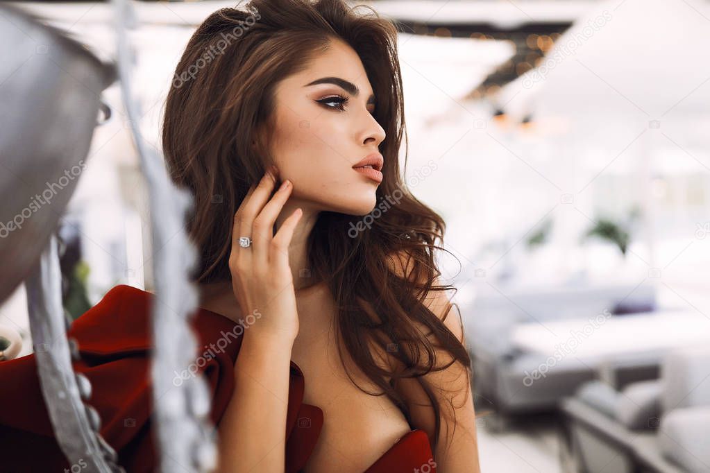 Fashion portrait of woman in red cocktail dress posing for modern magazine. Hand near the face, ring on the finger. Decollete and open shoulder. Dark long hairstyle. Standing in restaurant near vase