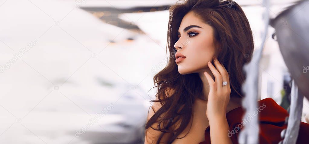 Gorgeous trendy girl standing in sexy res dress, perfect look, hand on the face, looking away. Long curly hair lying on the shoulder.Ring on the finer. Natural makeup.Metal designer vase with tentacle