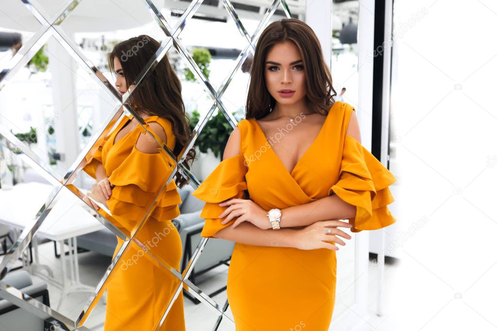 Passion woman wearing yellow evening dress with sleeves and decollete. Hands together, watches and neckless. Long dark hair lying o the back.Bright party makeup and fashion style. Reflecting in mirror