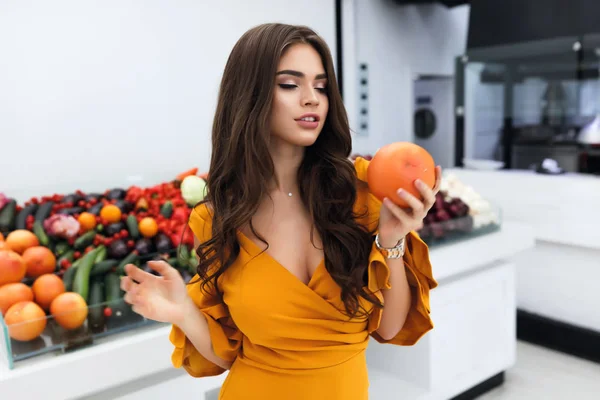 Attractive girl holding a grapefruit in a hand, smelling it and wants to try the taste. A lot of vegetables and fruits on a counter behind her. Perfect slim body, stylish hair and modern makeup.