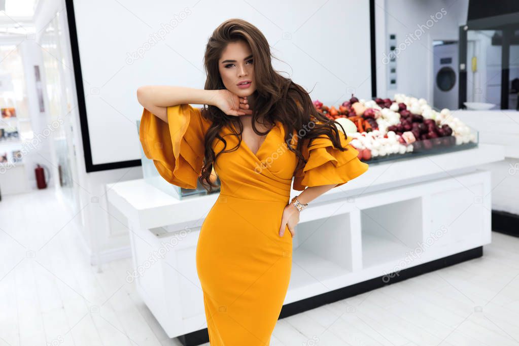 Gorgeous woman with volume brunette hairstyle, wearing evening cocktail yellow dress with sleeves and decollete. Party makeup on the face, big lips, white teeth. Standing near the vegetable counter.