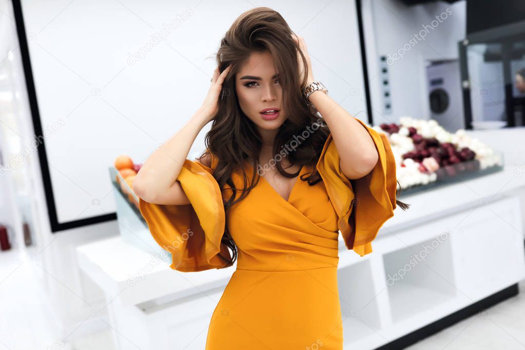 Portrait of stylish hot lady with long dark volume hair, hands on a head, bright makeup. Standing in shop on white background, vegetables and fruits behind her. Wearing fashion yellow dress.