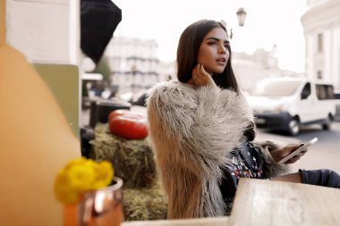 Modern genç kadın içinde café açık oturma, uzağa arıyorsunuz, smartphone elinde tutan kabarık gri ceket. Moda sonbahar rahat bakmak. Ahşap sehpa ile bir vazoda sarı çiçekler.