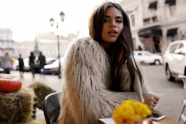 Moda sonbaharda açık çekim güzel esmer model bak. Kadının yüzündeki çıplak makyaj ile portre. Uzun kıvırcık saç omuz üzerine düşme. Kabarık ceket ve telefon. Saman ve kabak arkasında
