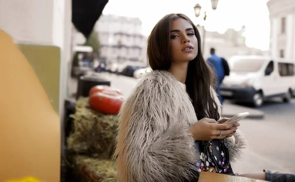 Glamorous lady with cell phone in hands talking to someone in internet. Black purse with flowers is on her knees. Long dark hair lying on shoulder, stylish makeup, big lips.Pumpkins on, hay background