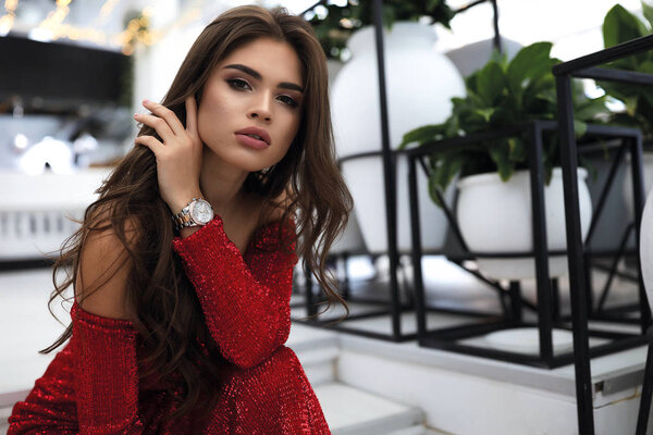 Fashion portrait of young brunette in red evening dress, accessories. Hand near the face, nude makeup.Long dark hair falling down on shoulders. Sitting on the steps of restaurant.Big lips, tender skin