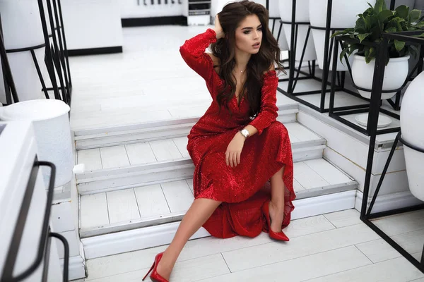 优雅性感的模特穿着红色的时装礼服坐在台阶上 高跟鞋在脚上 现代化妆和发型 长发躺在胸前 望向外 手放在头上 室内拍摄 — 图库照片