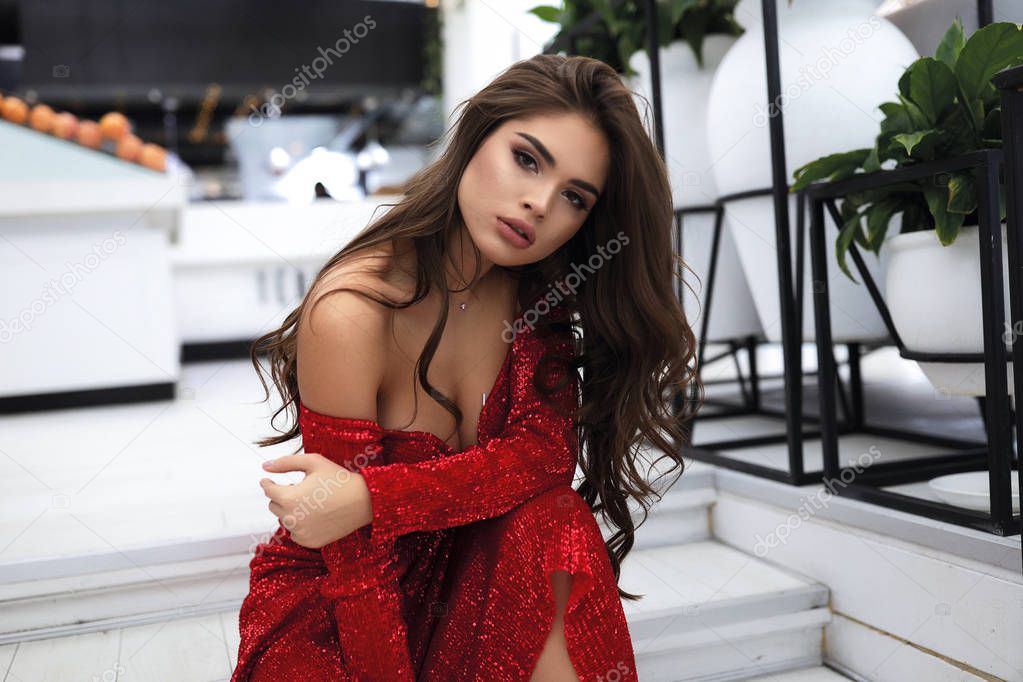 Lovely girl sitting on steps in luxury restaurant looking forward. Wearing red evening dress with decollete and open shoulders.Long curly hairstyle and modern makeup. Hand on arm.Fruits on background 