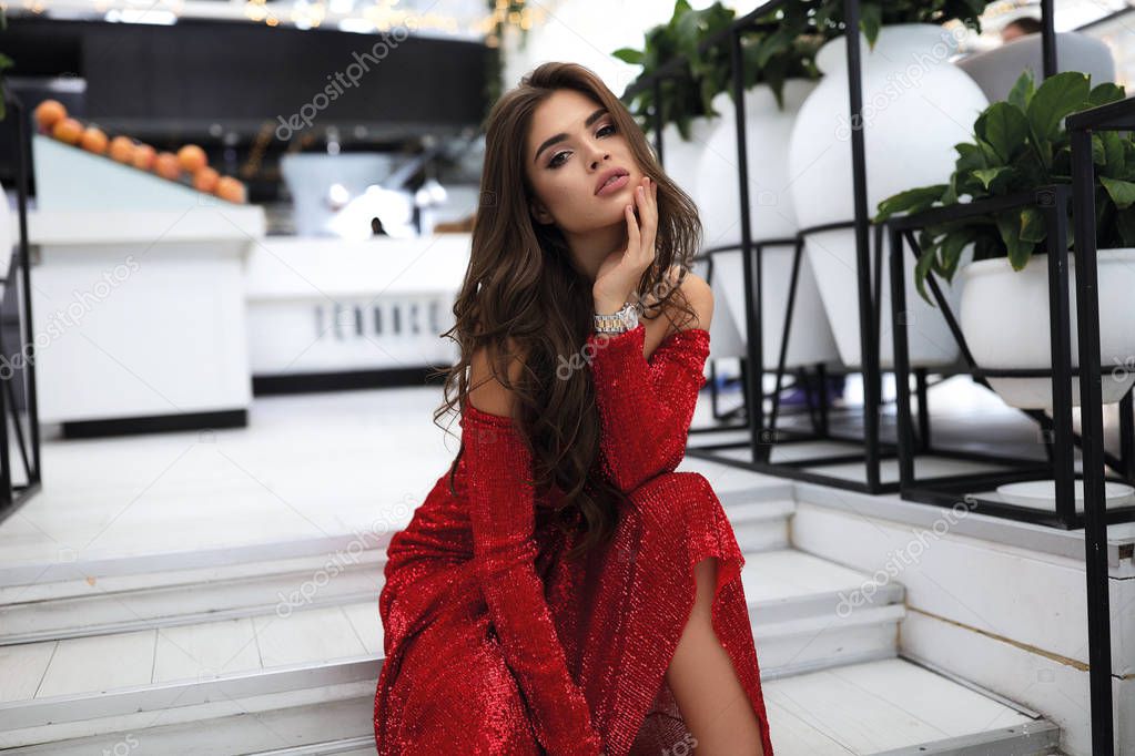 Passion model with long curly hairstyle and bright evening makeup. Sitting on the white steps in restaurant,wearing  red long party dress with open shoulders, watches on hand. Stylish fashion look