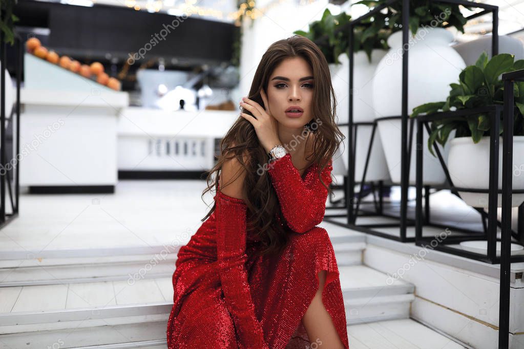 Effective bright lady in red long dress, sitting on white steps of the modern restaurant. Makeup on face, stylish curly hair lying on shoulders. Prepared for the party. Accessories, big lips, sparkles