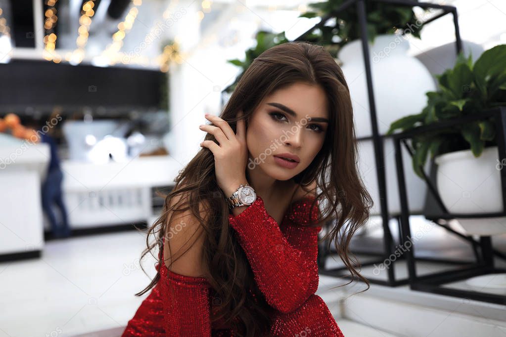 Beauty portrait of tender lady in red party dress with open shoulders. Tender hand touching face, modern watches on wrist. Brunette curly hairstyle and bright evening makeup.White design on background