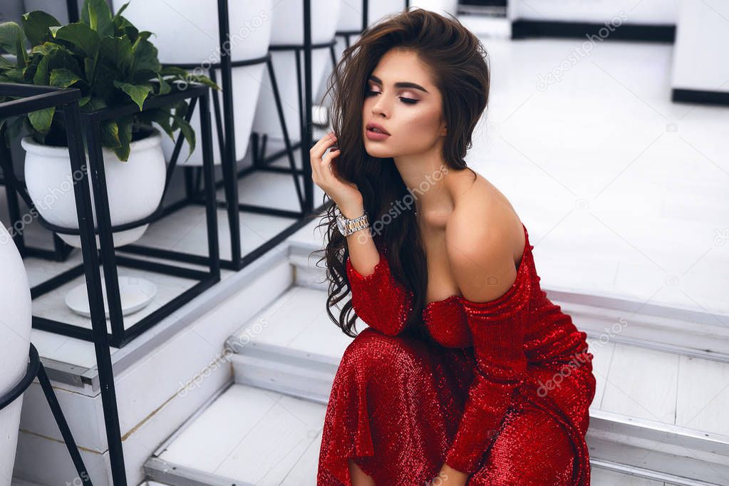 Sweet girl with shapely slim body wearing long red dress for an event. Open shoulders, hand near hair, big lips, accessories. Bright evening makeup and hairstyle. Sparkles on dress. Indoor shooting