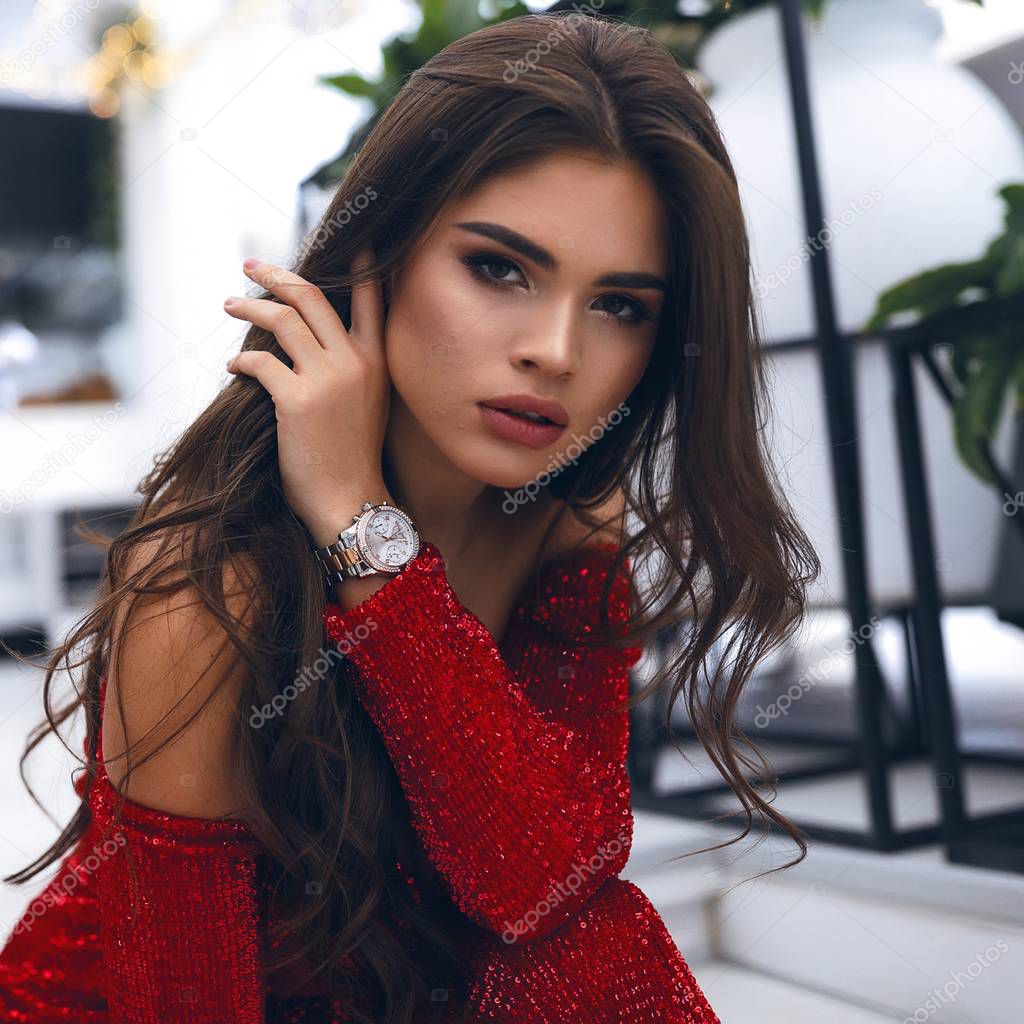 Close-up portrait of tender female with brunette long hair, dark eyes, big lips and nude makeup. Hand with watch touching hair, looking forward. wearing red party dress with sparkles, open shoulders.