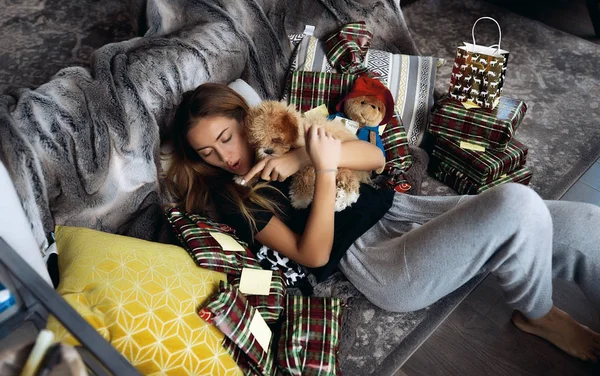 グレーのソファの上の彼女の小さな子犬と遊ぶ若い美しい女性 居心地の良い家庭的な雰囲気で 多くの柔らかい枕 小さな黒い鼻と赤巻き毛犬 親戚や友人にクリスマス プレゼント — ストック写真