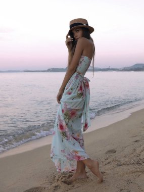 Yaz koleksiyonu sahilde poz ince genç model Giydir. Pembe çiçekler ve kemer ile hafif beyaz elbise göstermektedir. Hasır şapka kafasına kenarına el ile tutarak. Günbatımı 