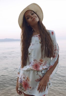 Gülen ve having fun Okyanusu yakınlarındaki sahilde neşeli bayan ayakta. Moda yaz bak, parlak çiçek ve şapka ile beyaz kısa elbise giyiyor. Uzun esmer saç göğüs yalan.