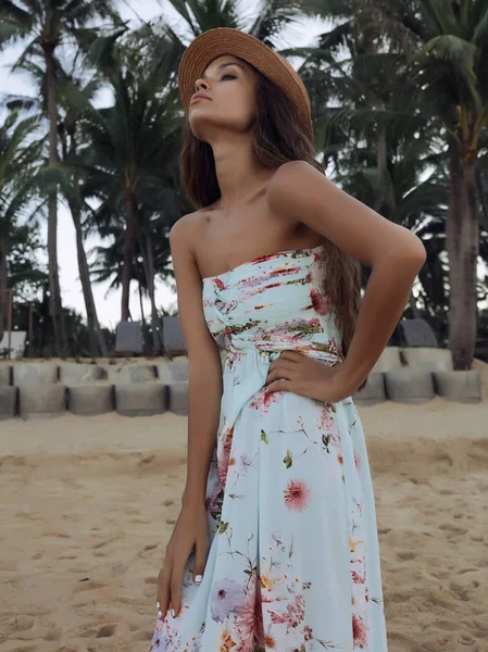 迷人的黑发女孩 长卷发 头上戴帽子 夏天的礼服与鲜花站在沙滩上 望着平静广阔的海洋 背景上的绿色高棕榈树 — 图库照片