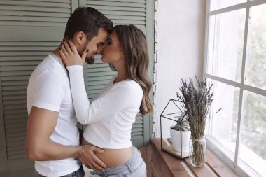 Güzel çift ayakta hafif pencere ve yeşil Panjur yakın. Sarılma ve öpüşme. Dokunaklı kadının karnında genç adam. Koca burnunu öpüşme hamile kadının yüzünde ver. Makyaj ve saç
