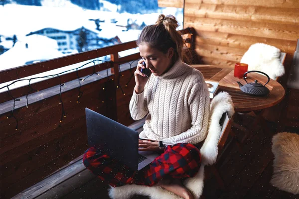 Pretty woman doing her job on computer online in the internet, talking on phone with the customer. Laptop lying on her knees, smart watches on the wrist. Comfortable home clothes, pajama and sweater