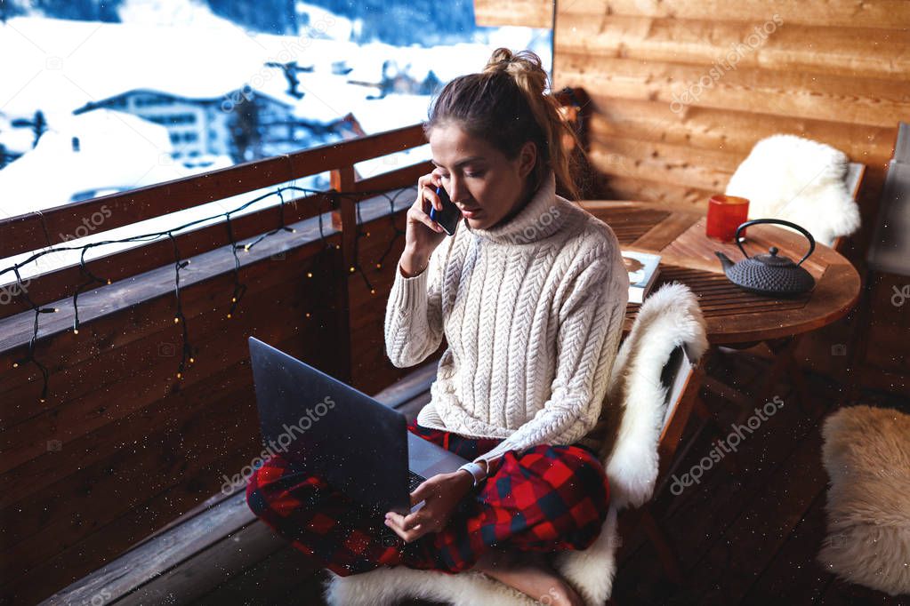 Lovely intelligent woman working remote on laptop and talking on phone. Searching the information in the internet, communicate with partners. Home comfortable style. Oversize sweater and red pants