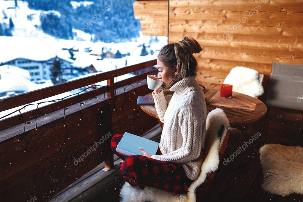 Blonde with long hair and oversize sweater sitting on the bench covered with natural sheep fur, holding her favourite book and drinking tasty coffee with amazing aroma. Wooden furniture and walls.