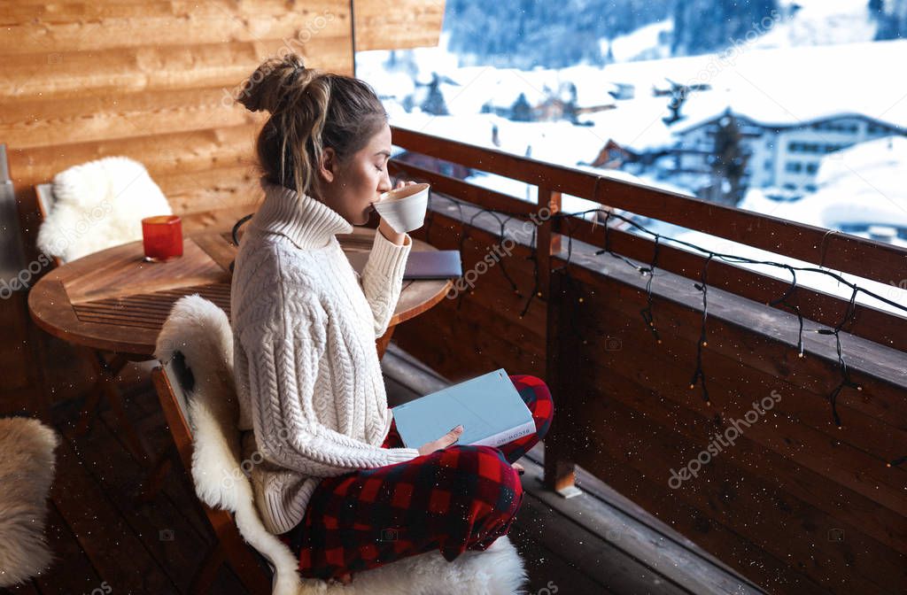 European morning with amazing winter view on the houses and mountains. A lot of white snow, pine trees around. Peaceful lady closed her eyes and enjoys the most aromatic coffee in the world. Reading