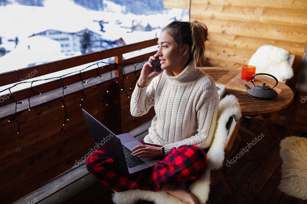 Serious and responsible girl talking on the phone about her work. Laptop lying on her knees, she working remote. Wooden coffee table with teapot and cups. Soft natural sheep fur on the chair.