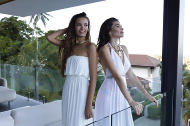 Neşeli esmerler şaşırtıcı deniz ile Balkonda duran görüntüleyin ve yaz tatillerini sahiptir. Modern ev, yüksek yeşil palmiye ağaçları. Beyaz ışık ince organları üzerinde elbise. Omuz üzerinde uzun süre ıslak saç