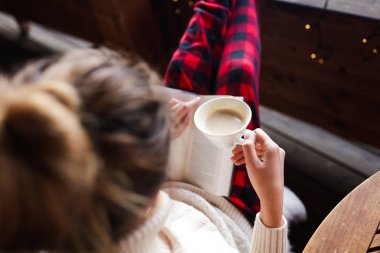 İhale ile sabah aromatik fincan kahve ve bir kitap terasta oturan Bayan okuma ve kış tatil sahiptir. Kırmızı kareli pantolon ve krem kazak. Topuz saç modeli. Taze dağ hava