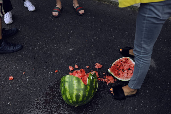Вкусный красный арбуз, лежащий на земле, разбился на две части. Маленькие кусочки разбросаны. Люди стоят вокруг него и смотрят. Женские ноги в джинсах. Летний отдых

