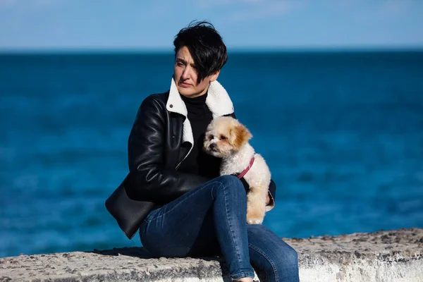 海の近くの桟橋に座っている黒いジャケットとジーンズのスタイリッシュな女性 巻き毛の毛皮で彼女のかわいい子犬を保持し 新鮮な空気でそよ風 屋外を歩く晴れた暖かい日 — ストック写真