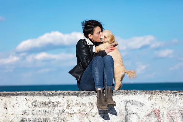 陽気な若い女性は 彼女の小さな子犬と桟橋で楽しんで キスし 彼を抱きしめています 晴れた春の天気 背景に青い空と波 革のジャケットとジーンズを着ている女性 — ストック写真