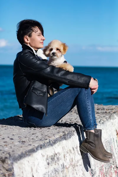 スタイリッシュなショートヘアとファッショナブルなスキニージーンズ レザージャケットを持つ夢の女性 犬と一緒に海の桟橋に座って 小さな友人が女性のひざの上に横たわっていた 屋外散歩のための晴れた日 — ストック写真