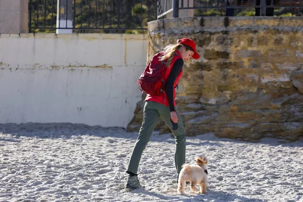 美しい若いブロンドは 小さなカーリー子犬と屋外を歩いています 海の近くの砂浜で楽しんでいるかわいい犬 赤いジャケット カーキパンツ バックパック 赤い帽子をかぶり リードを持つ女性 — ストック写真