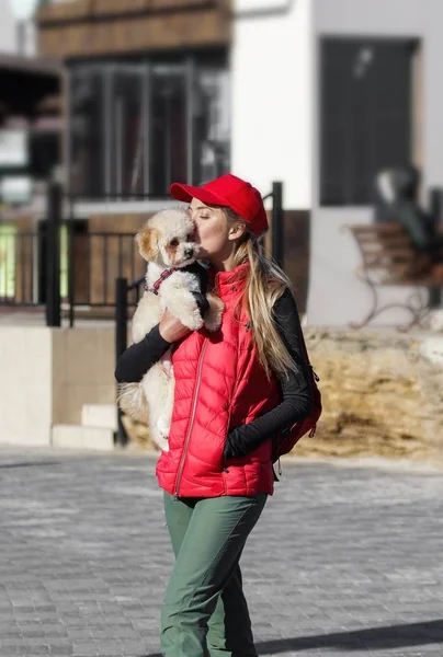 Cheerful and happy young woman holding her cute puppy on her hands and kissing him in a face. Small fluffy dog walking on a sunny day outdoor. Girl wearing red cap, jacket and pants.