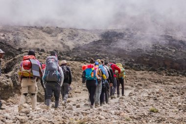 Afrika'ya ekstrem bir tur, Kilimanjaro'nun tepesine tırmanıyor. Bulutlu bir gün, yolda yürüyüş, ağır ekipman ve sırt çantaları çadır. Rahat spor giyim, güneş ışınlarından şapka. Kayalık zemin.