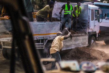 Sokakta çalışan, yol yapan çalışkan adamlar. Asfalt ile delik kazma ve doldurma. Sıcak Afrika havası, güvenlik için parlak yelekler. Zeminile büyük kamyon, bina için ekipman.
